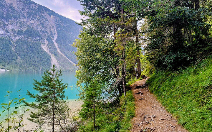Blog-Spend-based_Weight-based Ein Feldweg schlängelt sich durch üppig grüne Bäume am Ufer eines klaren, türkisfarbenen Sees entlang, während sich im Hintergrund unter einem teilweise bewölkten Himmel bewaldete Berge erheben.