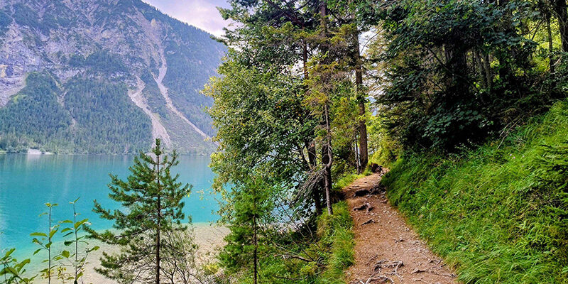 Blog-Spend-based_Weight-based Ein Feldweg schlängelt sich durch üppig grüne Bäume am Ufer eines klaren, türkisfarbenen Sees entlang, während sich im Hintergrund unter einem teilweise bewölkten Himmel bewaldete Berge erheben.