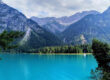 Ein türkisfarbener See mit klarem Wasser, umgeben von dichten Kiefernwäldern und hohen, schroffen Bergen unter einem teilweise bewölkten Himmel. Kiefernzweige rahmen den Blick im Vordergrund ein.