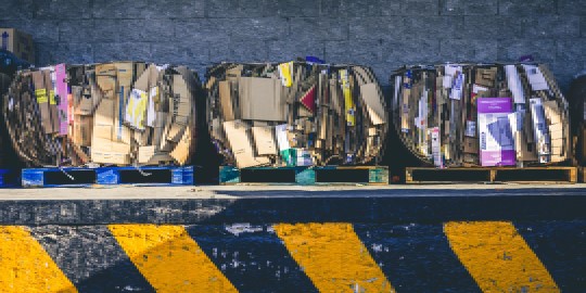Große Ballen aus gepresster Pappe und Papier, die mit Lansinoh SBT-Etiketten gekennzeichnet sind, stapeln sich auf Paletten vor einer Betonwand und einem gelb-schwarz gestreiften Bordstein.