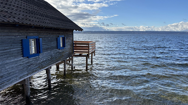 Ein Holzhaus mit blau umrandeten Fenstern steht auf Stelzen über dem klaren, plätschernden Wasser, neben einem Holzdeck mit Blick auf einen See unter einem teilweise bewölkten blauen Himmel - eine ruhige Szene, die an die COP 30-Ergebnisse für Klimaresilienz erinnert.
