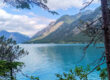 Ein ruhiger blauer See, umgeben von bewaldeten Bergen, mit einem kleinen weißen Segelboot in der Ferne unter einem teilweise bewölkten Himmel. Bäume rahmen den Vordergrund ein.