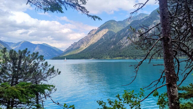 CRCF-Blogpost Ein ruhiger blauer See, umgeben von bewaldeten Bergen, mit einem kleinen weißen Segelboot in der Ferne unter einem teilweise bewölkten Himmel. Bäume rahmen den Vordergrund ein.
