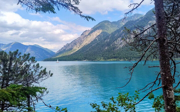 CRCF-Blogpost Ein ruhiger blauer See, umgeben von bewaldeten Bergen, mit einem kleinen weißen Segelboot in der Ferne unter einem teilweise bewölkten Himmel. Bäume rahmen den Vordergrund ein.