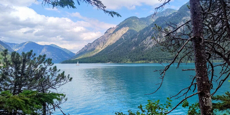 CRCF-Blogpost Ein ruhiger blauer See, umgeben von bewaldeten Bergen, mit einem kleinen weißen Segelboot in der Ferne unter einem teilweise bewölkten Himmel. Bäume rahmen den Vordergrund ein.