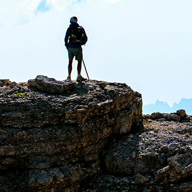 A hiker with a backpack and walking stick stands on a rocky cliff, overlooking distant mountain peaks under a hazy sky.