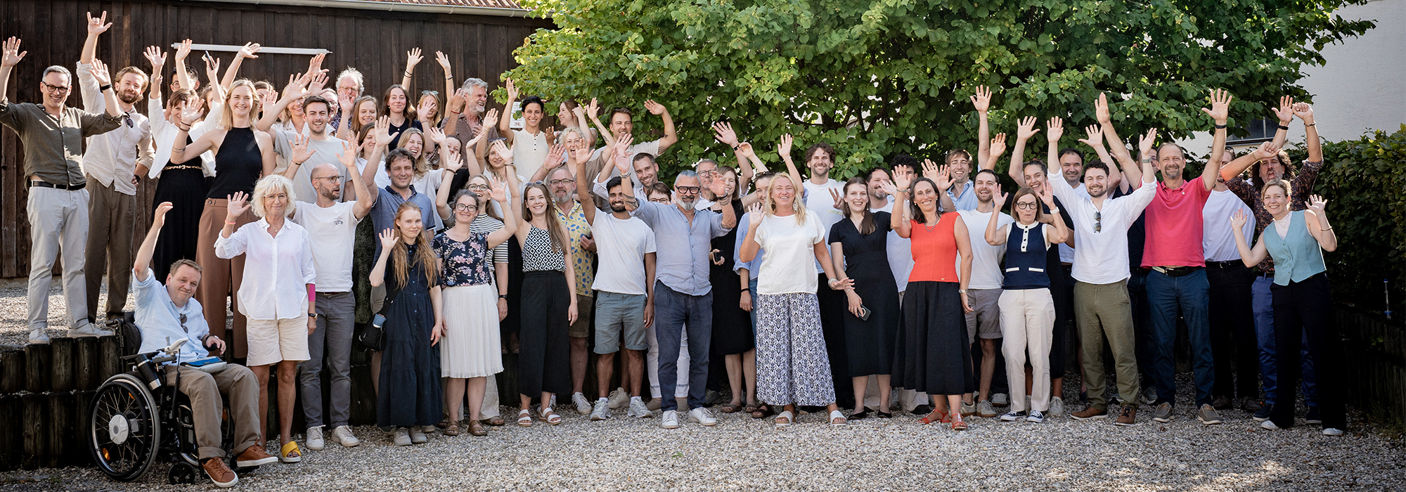 CertaniaSustainability-Team Eine große Gruppe von Menschen steht draußen auf Kies, lächelt und hebt begeistert die Hände. Im Hintergrund ist ein Baum zu sehen, und vorne links sitzt eine Person im Rollstuhl.