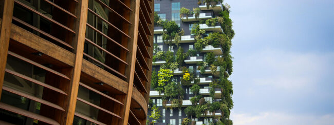 Corporate-Carbon-Footprint_Header A tall modern building with balconies full of lush green plants stands next to another building with wooden slats, under a partly cloudy sky.