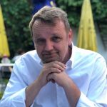 Dr_Fleissner_Profile_Image Ein Mann mit kurzen hellbraunen Haaren und einem Bart, der ein weißes Hemd trägt, sitzt im Freien, hat die Hände unter dem Kinn gefaltet und lächelt leicht. Im Hintergrund sind unscharfe gelbe Regenschirme und Grünpflanzen zu sehen.