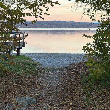 Ein Weg durch den Wald führt zu einem kleinen Kiesstrand und einem Holzsteg an einem ruhigen See bei Sonnenuntergang.