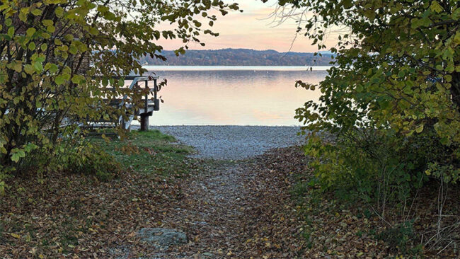 ESRS-2 Ein Weg durch den Wald führt zu einem kleinen Kiesstrand und einem Holzsteg an einem ruhigen See bei Sonnenuntergang.
