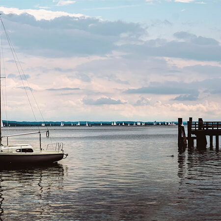 Ein Segelboot liegt in der Nähe eines hölzernen Stegs auf einem ruhigen See vor Anker, und auf dem Steg stehen Menschen. In der Ferne gleiten kleine Segelboote unter einem teilweise bewölkten Himmel dahin - eine idyllische Szene, die Harmonie widerspiegelt, ähnlich wie eine EcoVadis Bewertung.
