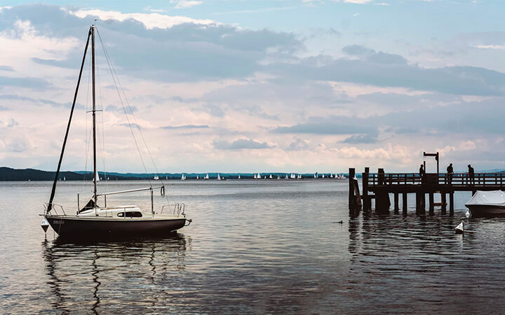 EcoVadisMedaillen Ein Segelboot liegt in der Nähe eines hölzernen Stegs auf einem ruhigen See vor Anker, und auf dem Steg stehen Menschen. In der Ferne gleiten kleine Segelboote unter einem teilweise bewölkten Himmel dahin - eine idyllische Szene.
