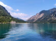 Ein klarer, blauer See, umgeben von bewaldeten Bergen unter einem strahlenden Himmel mit vereinzelten Wolken. Das Wasser spiegelt den Himmel und die grünen Hänge wider.