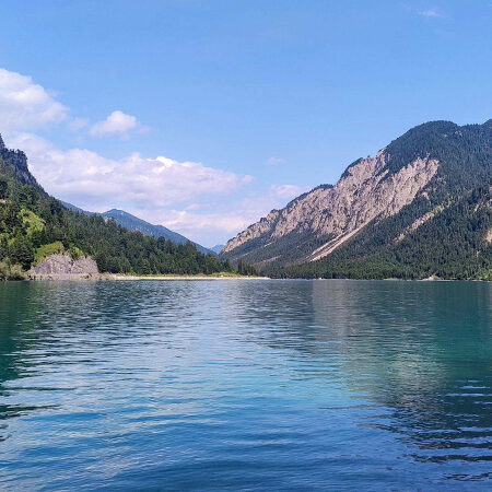 Ein klarer, blauer See, umgeben von bewaldeten Bergen unter einem strahlenden Himmel mit vereinzelten Wolken. Das Wasser spiegelt den Himmel und die grünen Hänge wider.