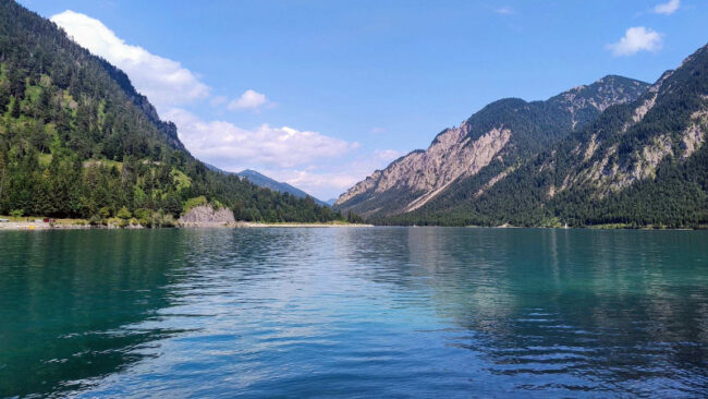 EmpCo-Richtlinie Ein klarer, blauer See, umgeben von bewaldeten Bergen unter einem strahlenden Himmel mit vereinzelten Wolken. Das Wasser spiegelt den Himmel und die grünen Hänge wider.