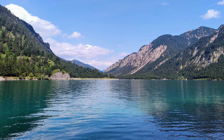 EmpCo-Richtlinie Ein klarer, blauer See, umgeben von bewaldeten Bergen unter einem strahlenden Himmel mit vereinzelten Wolken. Das Wasser spiegelt den Himmel und die grünen Hänge wider.