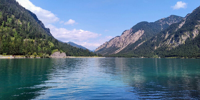 EmpCo-Richtlinie Ein klarer, blauer See, umgeben von bewaldeten Bergen unter einem strahlenden Himmel mit vereinzelten Wolken. Das Wasser spiegelt den Himmel und die grünen Hänge wider.