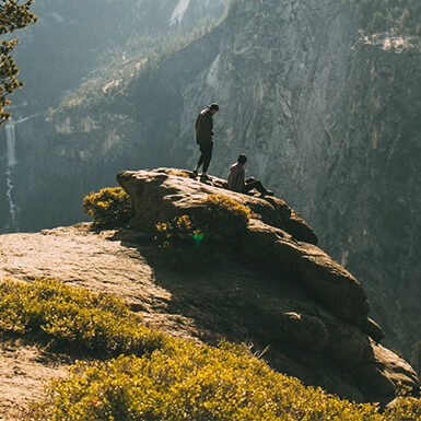 Two people stand and sit on a rocky cliff edge overlooking a deep valley with mountains and a distant waterfall, surrounded by trees and greenery in a scenic, natural landscape.