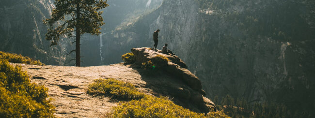Two people stand and sit on a rocky cliff edge overlooking a deep valley with mountains and a distant waterfall, surrounded by trees and greenery in a scenic, natural landscape.