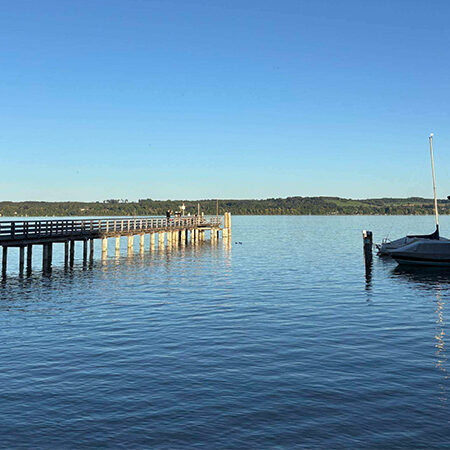 Ein langer Holzsteg, der in einen ruhigen See hineinragt, mit einem überdachten Bootshaus auf der rechten Seite.