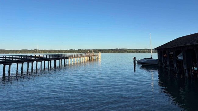 IOS1406 Ein langer Holzsteg, der in einen ruhigen See hineinragt, mit einem überdachten Bootshaus auf der rechten Seite.