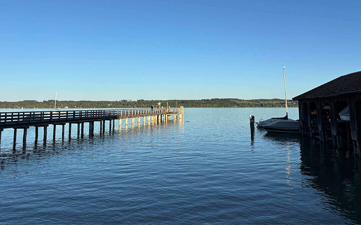 IOS1406 Ein langer Holzsteg, der in einen ruhigen See hineinragt, mit einem überdachten Bootshaus auf der rechten Seite.