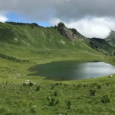 Ein kleiner, klarer See liegt inmitten eines grünen, grasbewachsenen Tals, umgeben von sanften Hügeln und felsigen Hängen unter einem teilweise bewölkten Himmel.