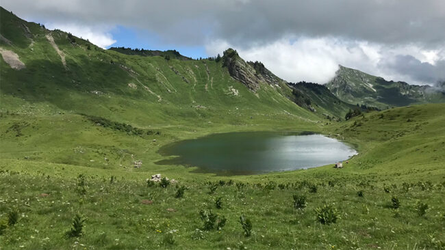 ISO-14001-2026 Ein kleiner, klarer See liegt inmitten eines grünen, grasbewachsenen Tals, umgeben von sanften Hügeln und felsigen Hängen unter einem teilweise bewölkten Himmel.