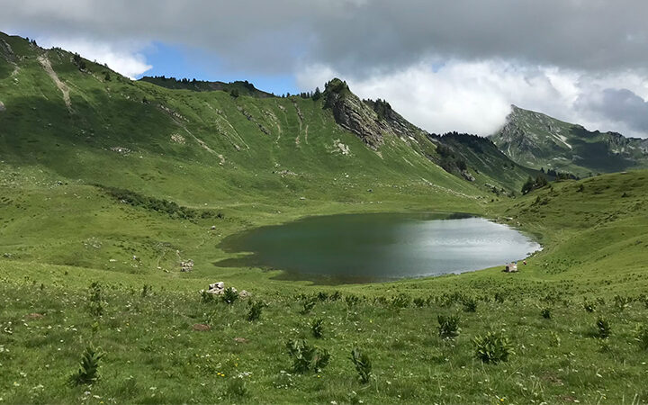 ISO-14001-2026 Ein kleiner, klarer See liegt inmitten eines grünen, grasbewachsenen Tals, umgeben von sanften Hügeln und felsigen Hängen unter einem teilweise bewölkten Himmel.