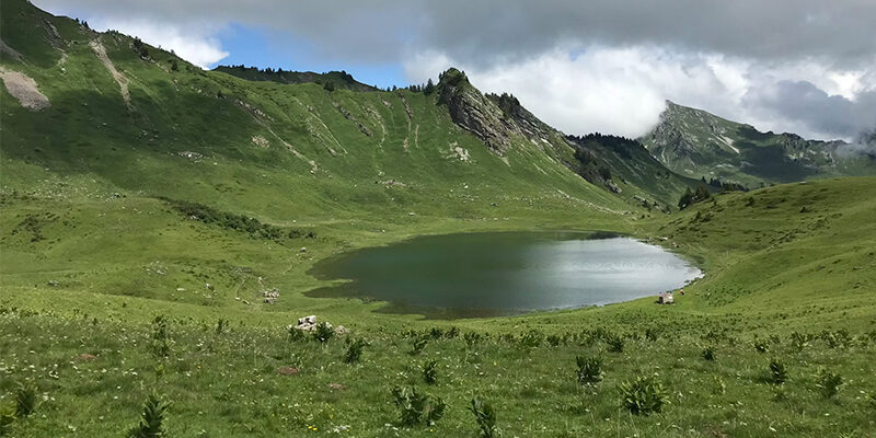 ISO-14001-2026 Ein kleiner, klarer See liegt inmitten eines grünen, grasbewachsenen Tals, umgeben von sanften Hügeln und felsigen Hängen unter einem teilweise bewölkten Himmel.