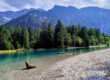 Ein klarer, blauer See mit Treibholz am felsigen Ufer, umgeben von dichtem, grünem Wald und hohen Bergen unter einem bewölkten Himmel.