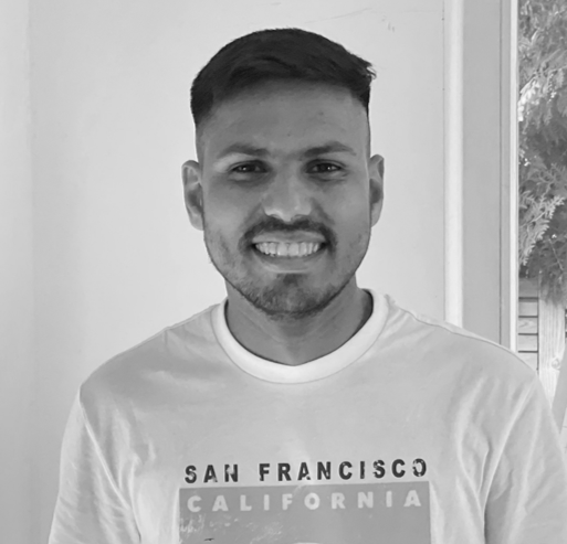 Ein junger Mann mit kurzen Haaren und einem Bart lächelt in die Kamera und trägt ein T-Shirt mit der Aufschrift San Francisco California. Im Hintergrund sind eine weiße Wand und ein Teil eines Fensters zu sehen, und das Bild zeigt die klassischen Schwarz-Weiß-Töne von Lansinoh SBT.