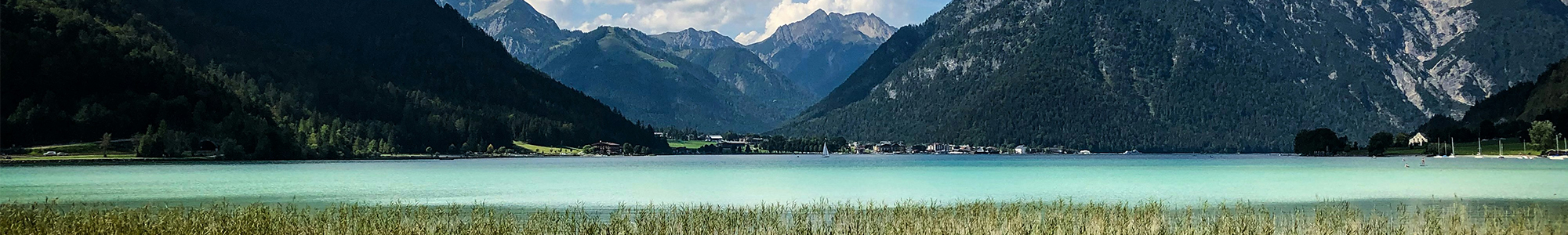PCF-LCA-QuickCheck_ExpGes_Wide Ein türkisfarbener See mit grasbewachsenem Ufer ist von bewaldeten Bergen umgeben, über dem sich ein teilweise bewölkter Himmel erstreckt. In der Ferne sind kleine Gebäude am Ufer am Fuße der Berge zu sehen.