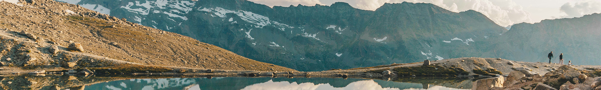 PPWR-Wide Zwei Menschen spazieren auf einem felsigen Pfad in der Nähe eines ruhigen Bergsees, im Hintergrund sind hohe Gipfel und Schneeflecken zu sehen, über ihnen ein teilweise bewölkter Himmel.