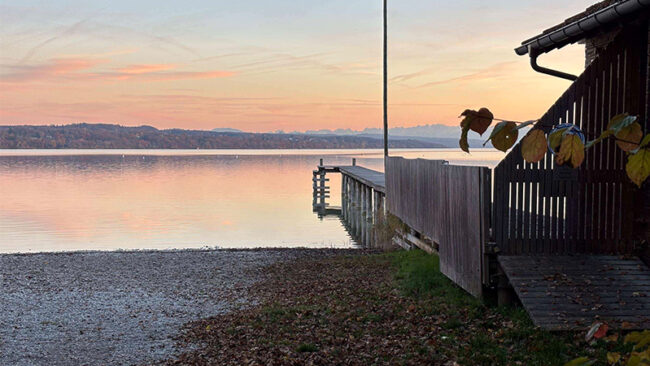 SBTi-Validierung Ein Holzsteg ragt bei Sonnenuntergang in einen ruhigen See hinein, betrachtet von einem Kiesweg neben einem Holzgebäude aus.