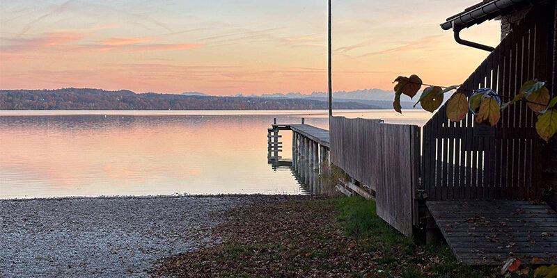 SBTi-Validierung Ein Holzsteg ragt bei Sonnenuntergang in einen ruhigen See hinein, betrachtet von einem Kiesweg neben einem Holzgebäude aus.