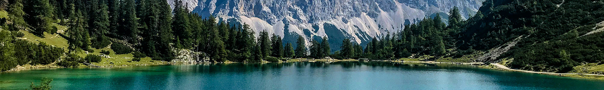 SBTiQuickCheck_Wide Ein klarer, türkisfarbener See, umgeben von Bäumen, mit hohen, felsigen Bergen im Hintergrund unter blauem Himmel.