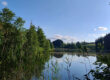 Ein ruhiger See, umgeben von hohem grünem Gras und dichten Bäumen, in dessen Wasser sich der blaue Himmel und vereinzelte Wolken spiegeln und so eine friedliche Naturkulisse schaffen.