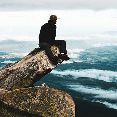 A person sits on a rocky outcrop overlooking a vast, mountainous landscape with clouds and distant peaks under a cloudy sky.