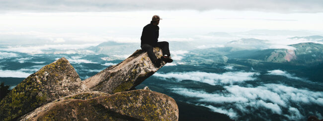 Eine Person sitzt auf einem Felsvorsprung mit Blick auf eine weite, bergige Landschaft, unter ihr liegen Wolken und Nebel; sie trägt einen Hut und dunkle Kleidung.