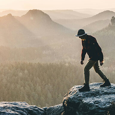A person wearing a hat walks on a rocky cliff at sunrise, overlooking a vast forest and distant hills bathed in soft, golden light.