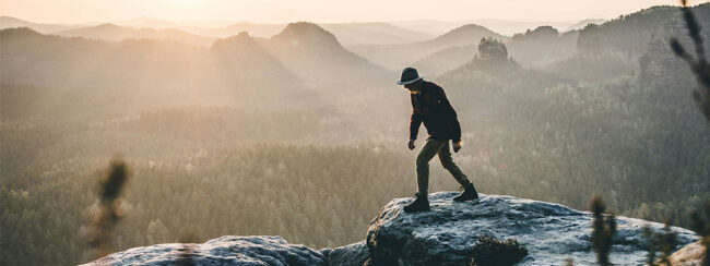 Eine Person mit Hut steht bei Sonnenaufgang auf einer felsigen Klippe und blickt auf einen weitläufigen, nebelverhangenen Wald und sanfte Hügel in der Ferne.