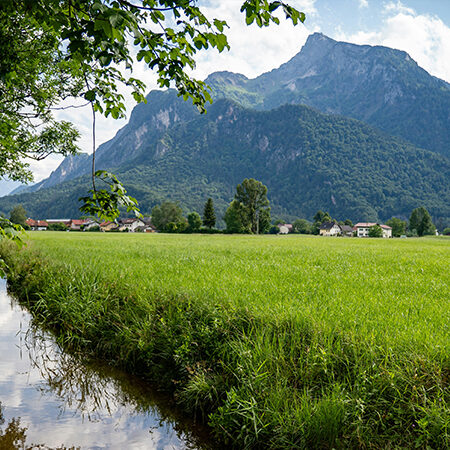 Ein schmaler Bach fließt entlang eines saftig grünen Feldes mit hohen, bewaldeten Bergen im Hintergrund unter einem teilweise bewölkten Himmel. In der Ferne sind Bäume und Häuser zu sehen.